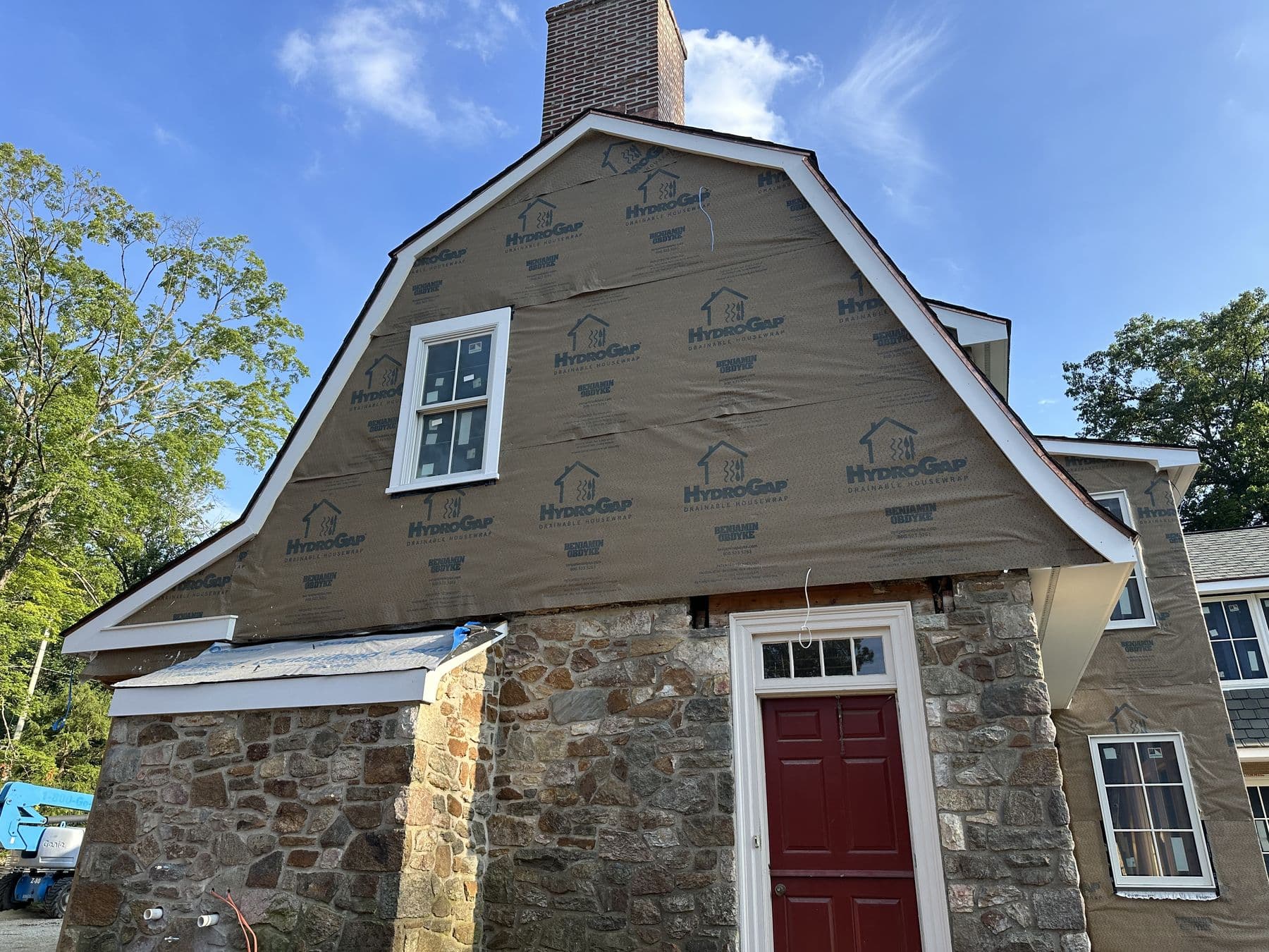 Gambrel end of the stone house with new windows and housewrap above a red door